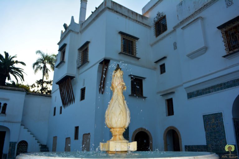 A whitewashed facade with a fountain urn beneath the palms.